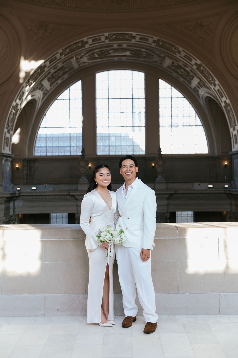 Couple in white outfits posing together under arched windows in historic building.