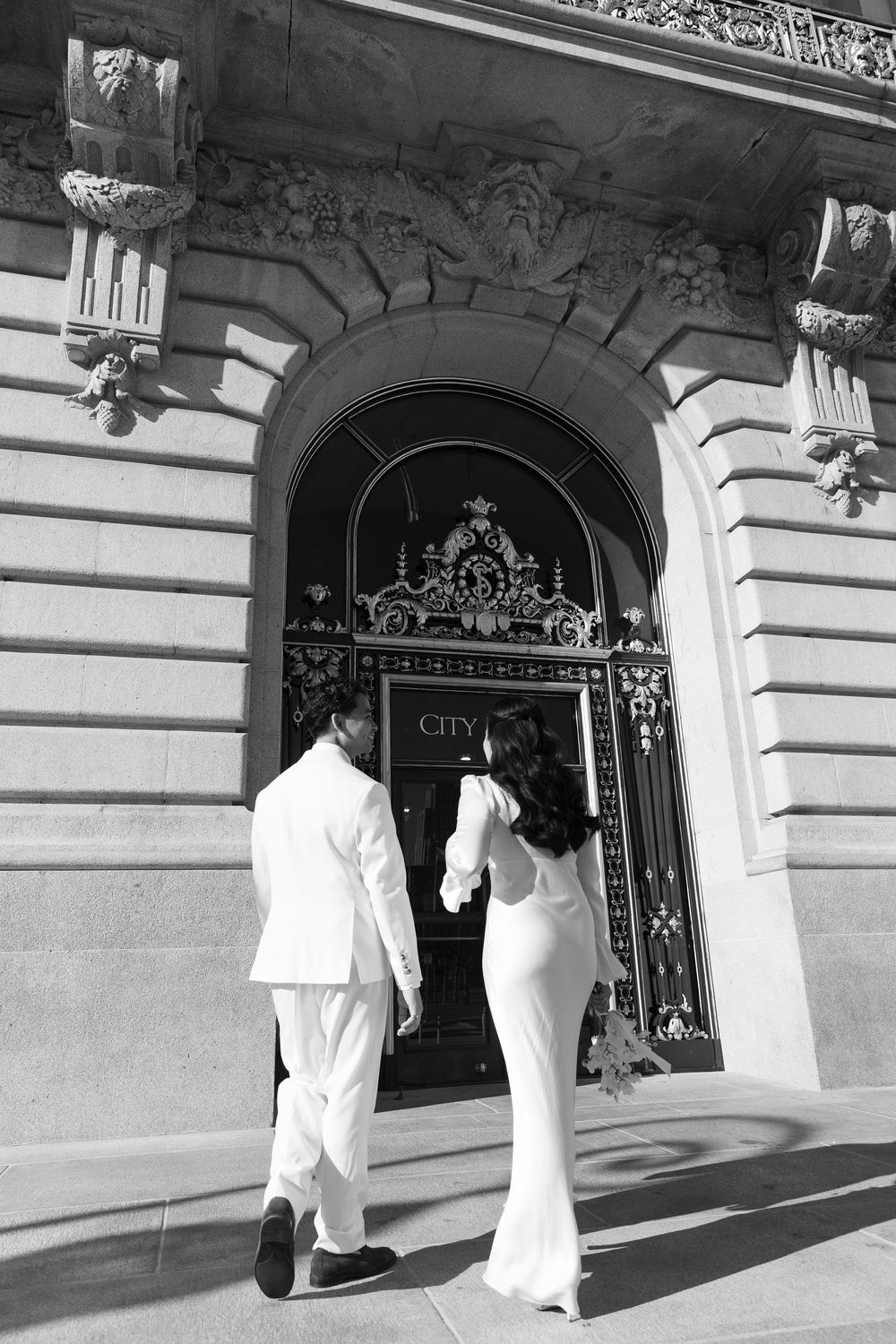 Couple in white attire entering through ornate City Hall doorway.