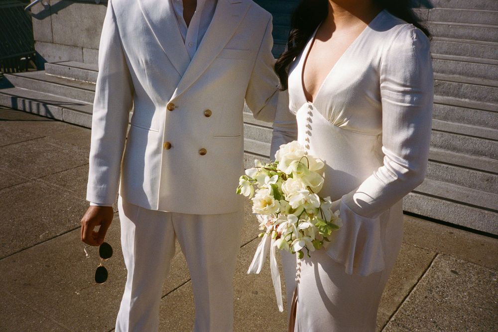 Cropped view of couple in white suits holding a bouquet of white flowers.