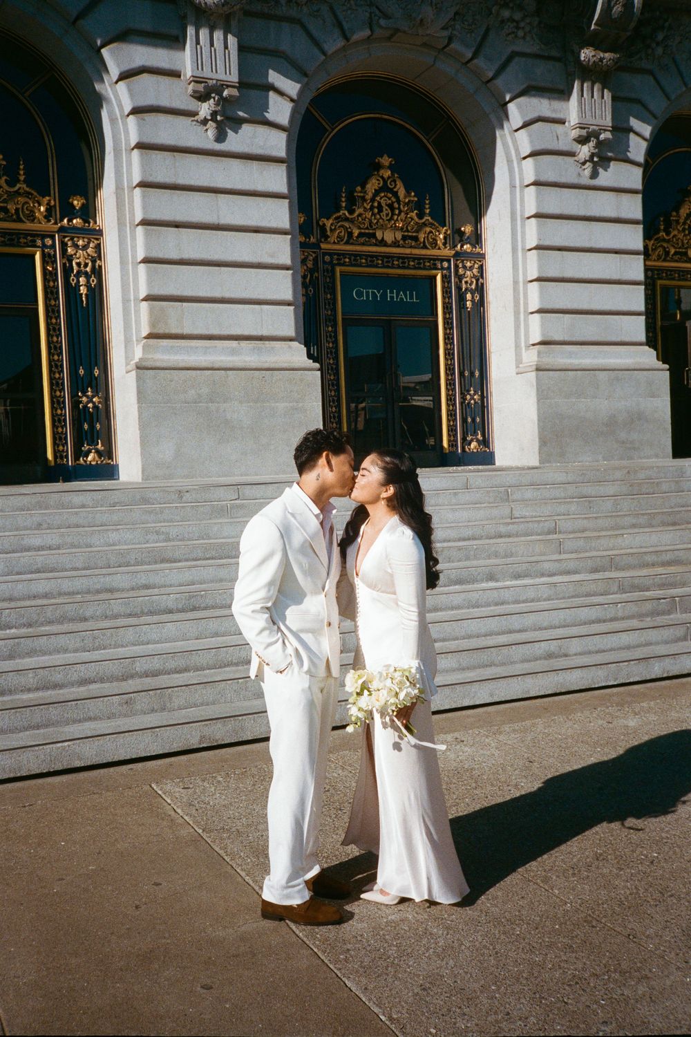 Wedding couple in white outfits posing outside historic City Hall building on stone steps.