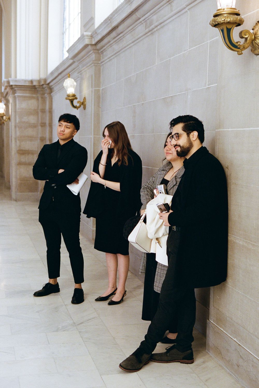Three people in dark formal attire stand against a light colored wall with sconces.