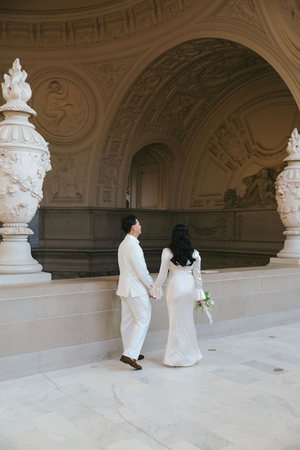 A couple in white outfits explores an ornate hallway with classical architecture and sculptures.