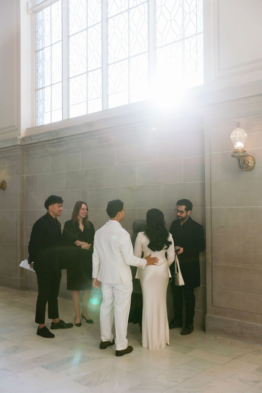 A small group gathers in an elegant room with high windows and stone walls wearing formal attire.
