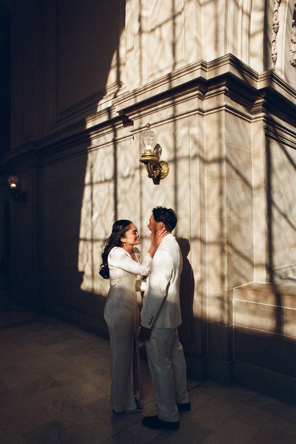 A couple shares an intimate moment against a sunlit stone wall with dramatic shadows.