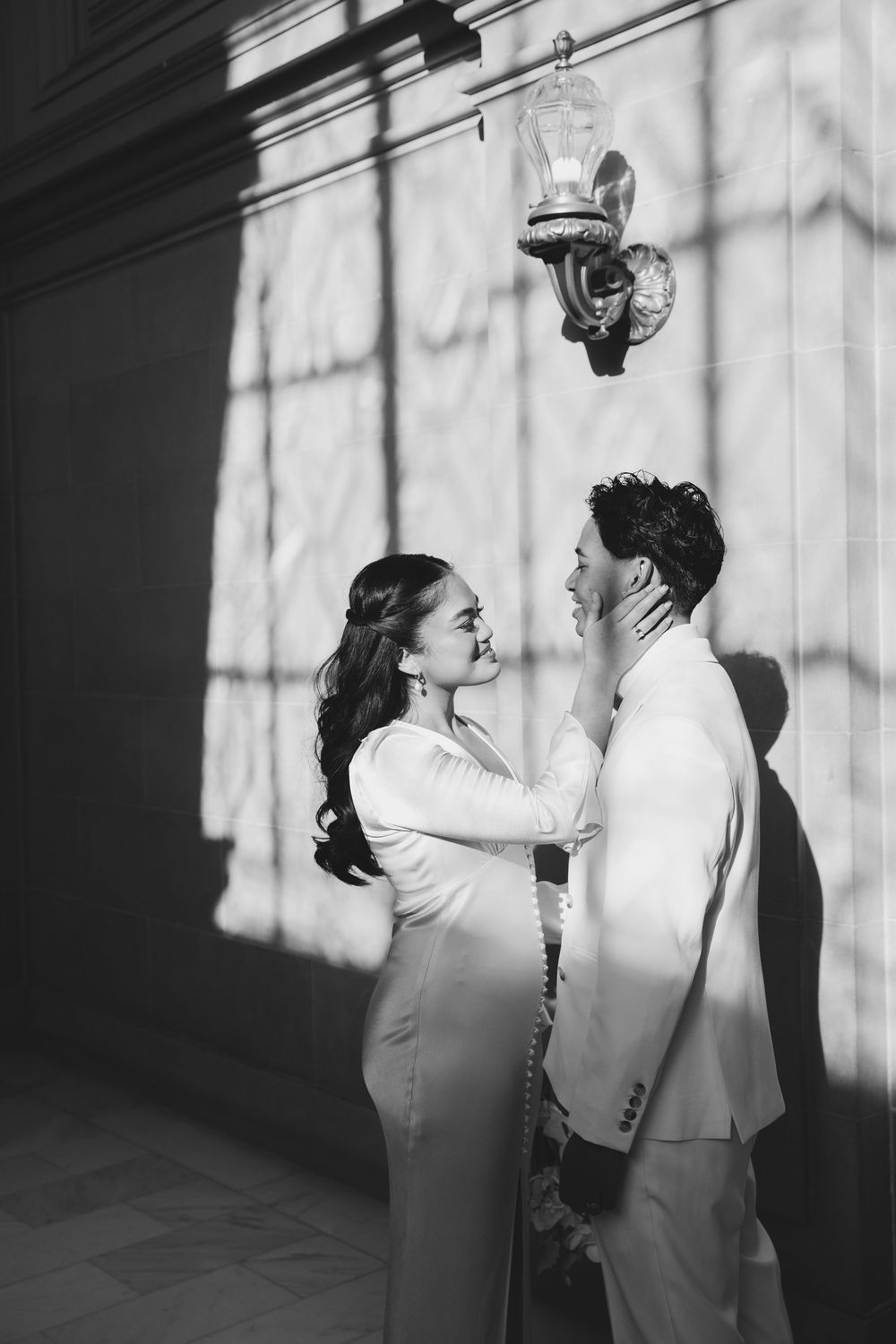 A black and white portrait of a couple embracing against a wall with dramatic window light.