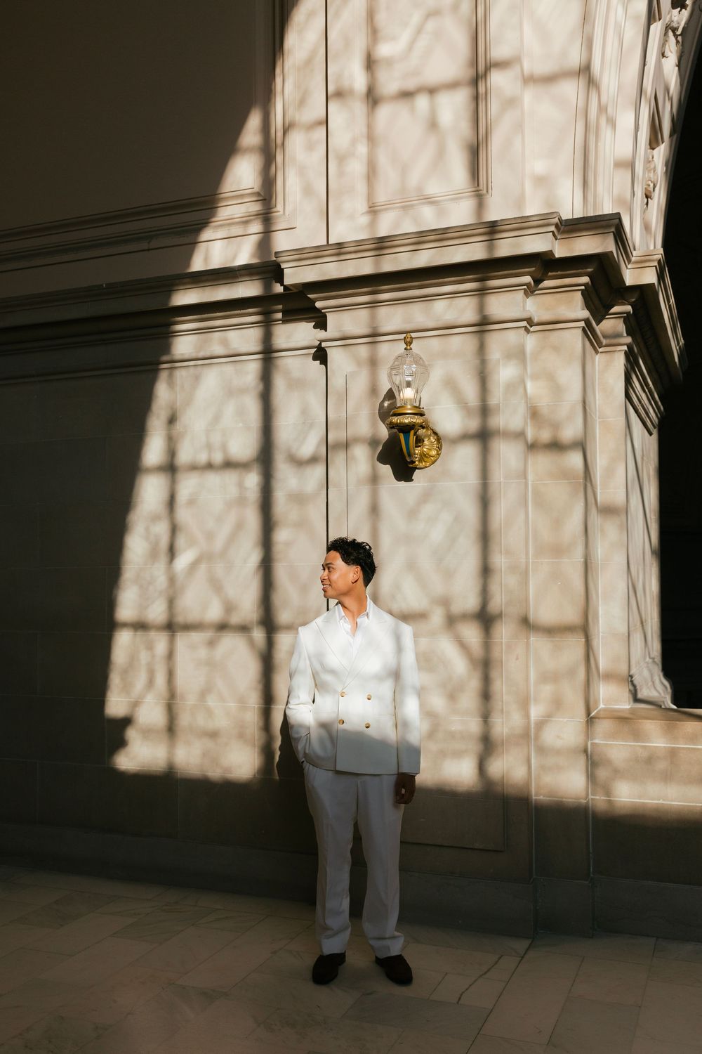 A person in a white suit stands alone against a wall with dramatic shadows and lighting.