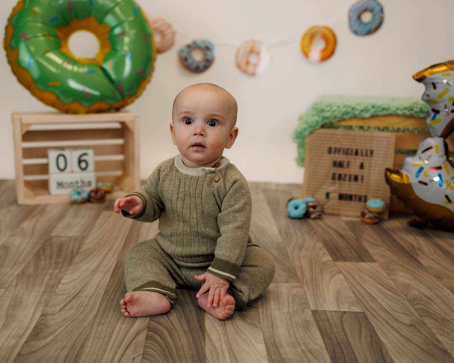 Smiling baby wearing coral sweatshirt and tiny gold crown sits in wooden barrel against beige background.