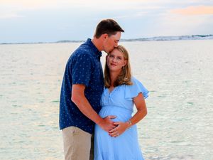A couple in blue clothing share a tender moment on the beach at sunset with calm ocean waters in the background.
