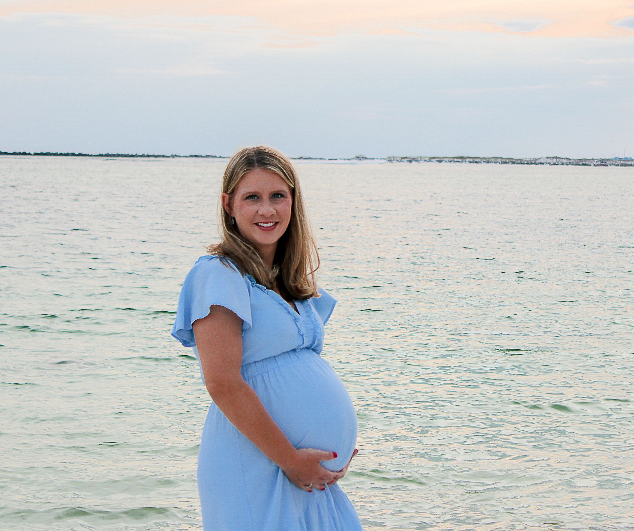 Someone wearing a light blue maternity dress stands by the ocean at sunset while cradling their baby bump.