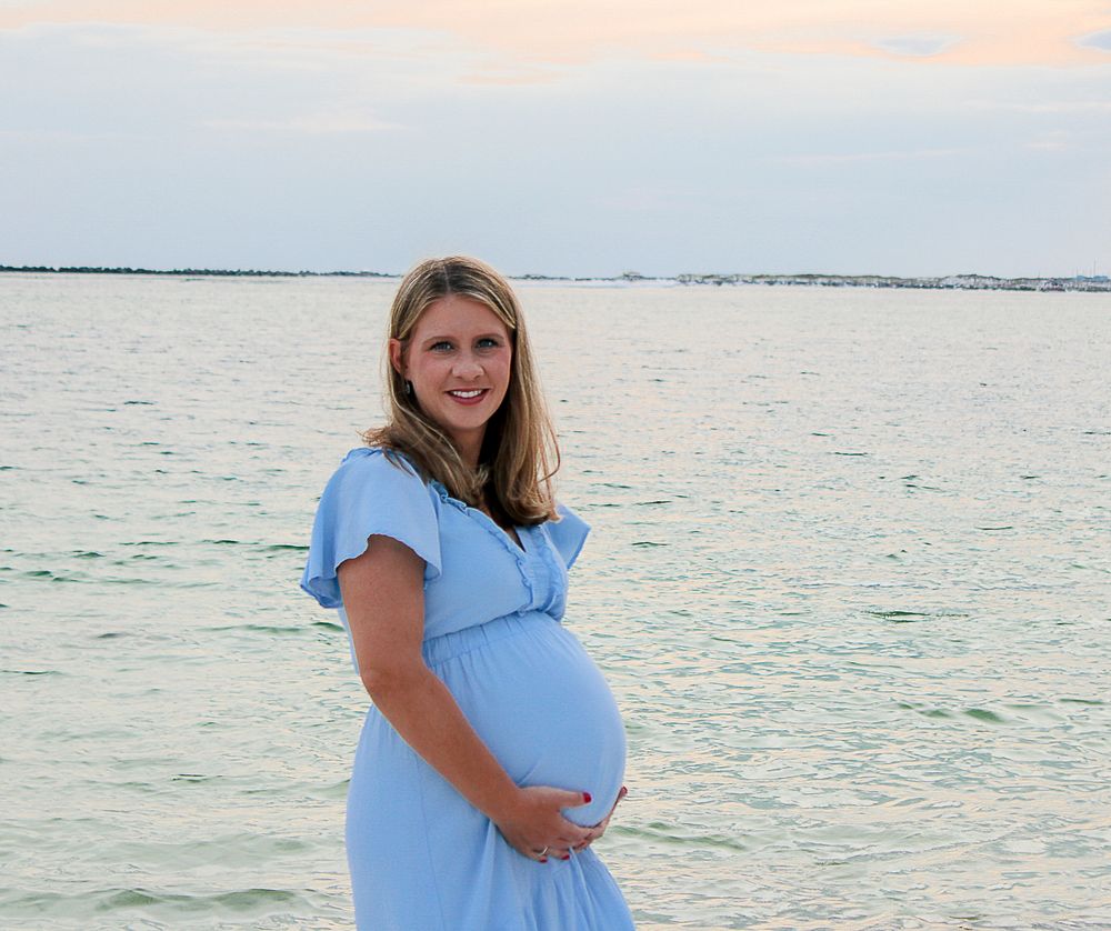 Someone wearing a light blue maternity dress stands by the ocean at sunset while cradling their baby bump.