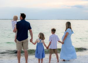 A family wearing light blue and navy clothing holds hands while looking out at the ocean during sunset.