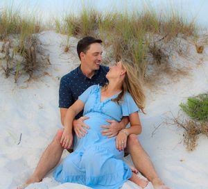 A couple shares a joyful moment sitting together on sand dunes with beach grass swaying in the background.