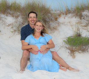Couple sits together on white sandy beach dunes with sea grass during maternity photo session at sunset.