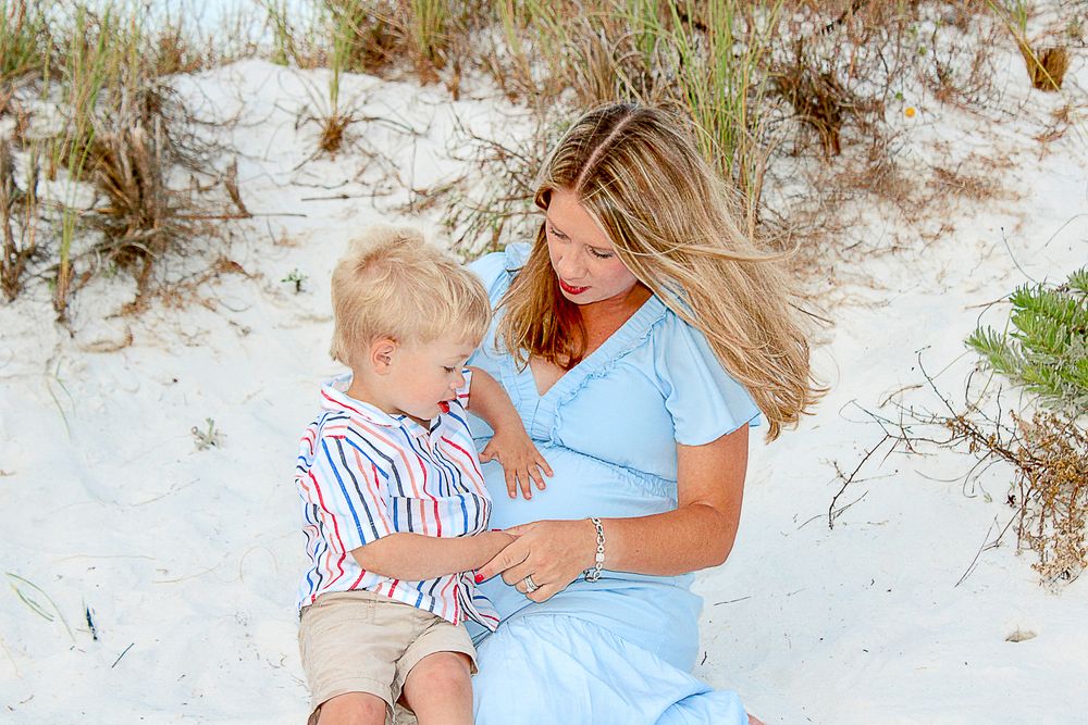 Parent and child sitting together on white sandy beach dunes wearing light summer clothing.