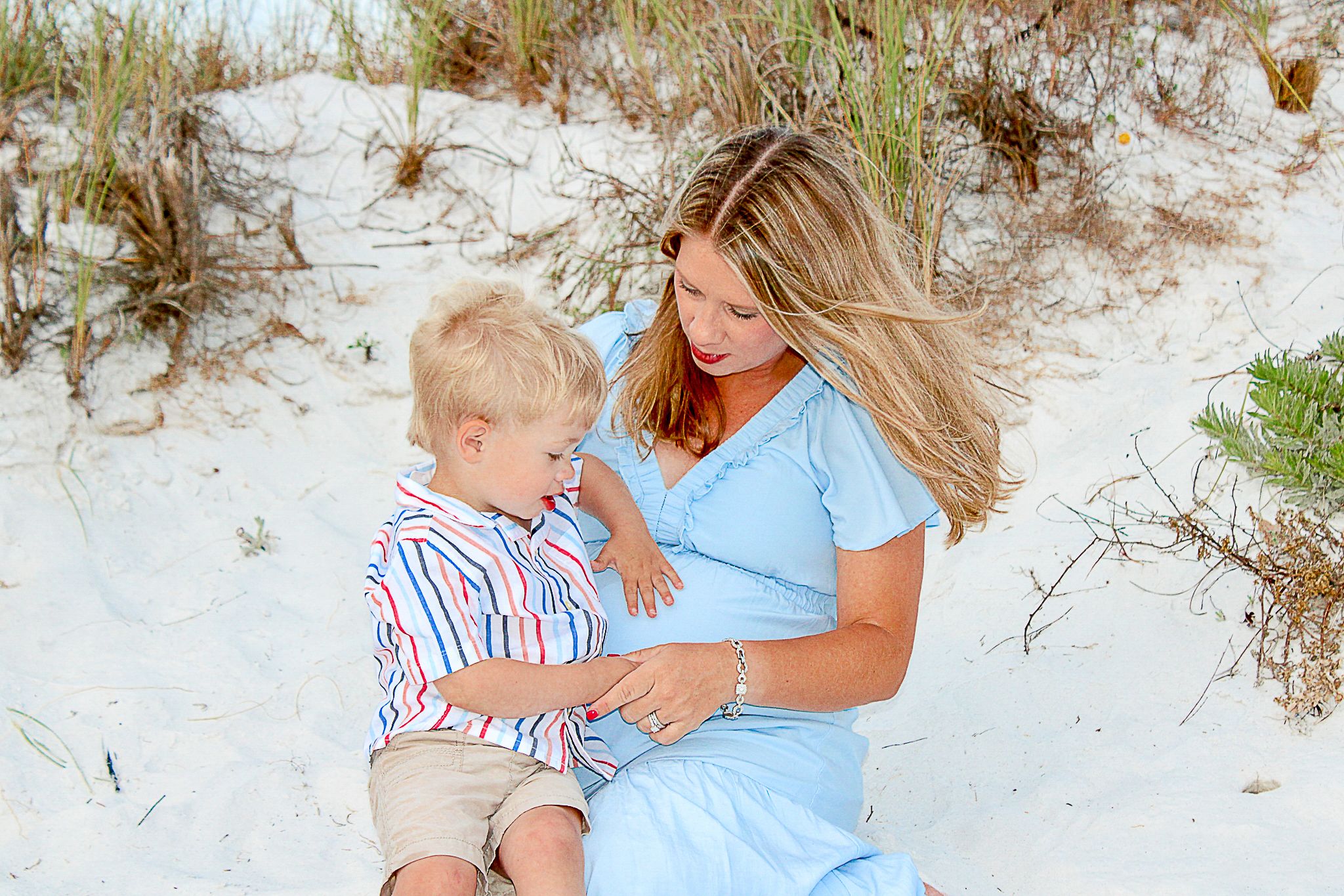 Parent and child sitting together on white sandy beach dunes wearing light summer clothing.