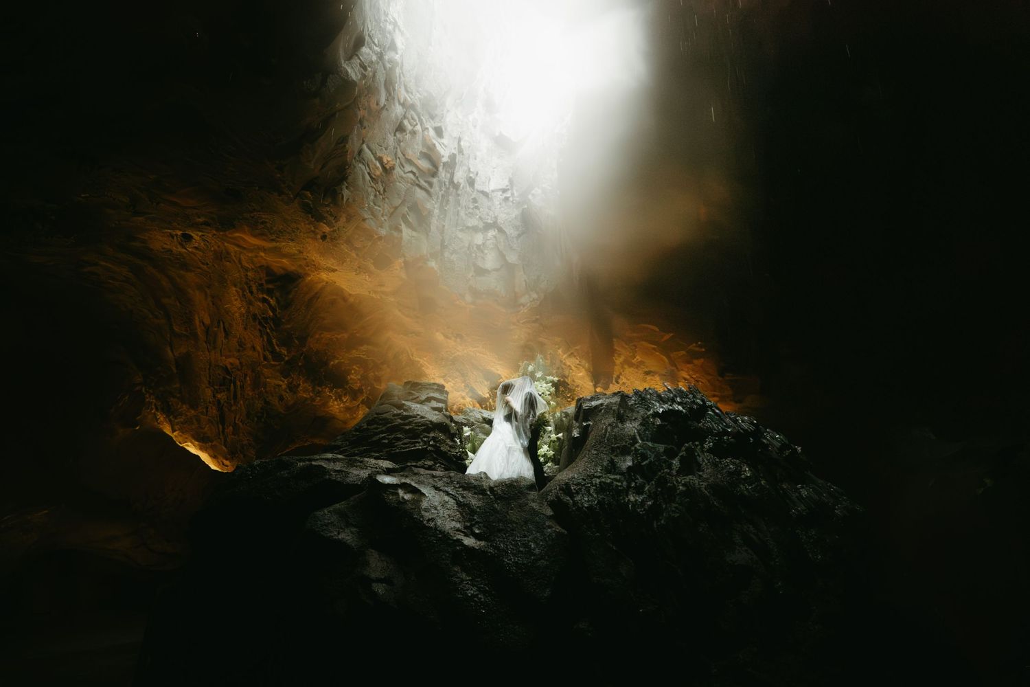 Dramatic cave wedding photo showing a couple on dark rocks illuminated by warm golden light filtering through from above.