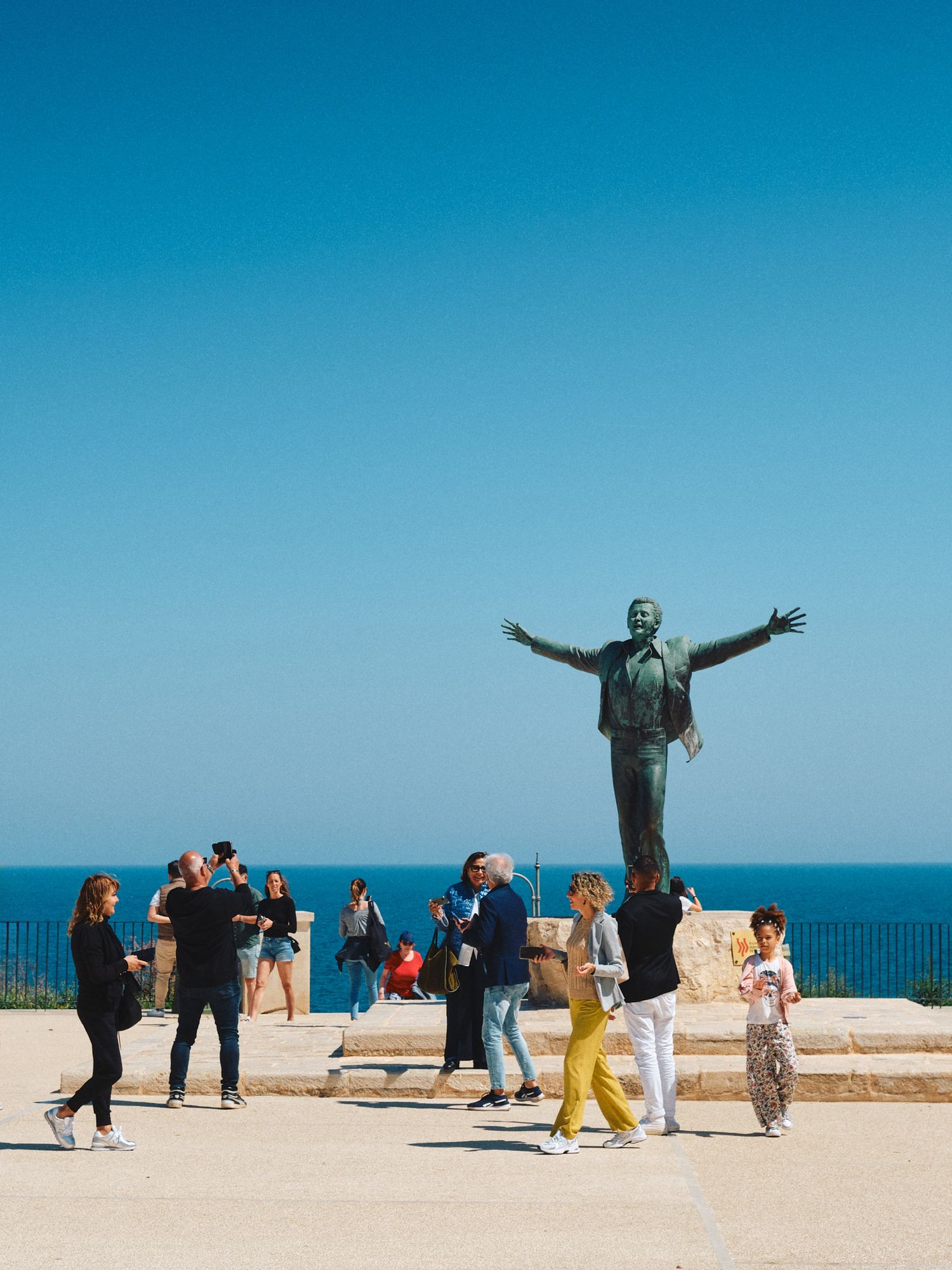 Tourists gather around a large statue overlooking the Mediterranean Sea on a bright sunny day with clear blue skies.