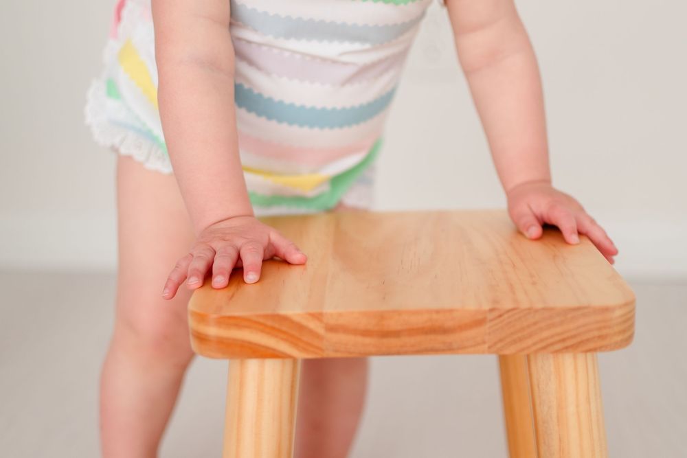 Small hands grip a wooden stool showing a toddler wearing pastel striped clothing.