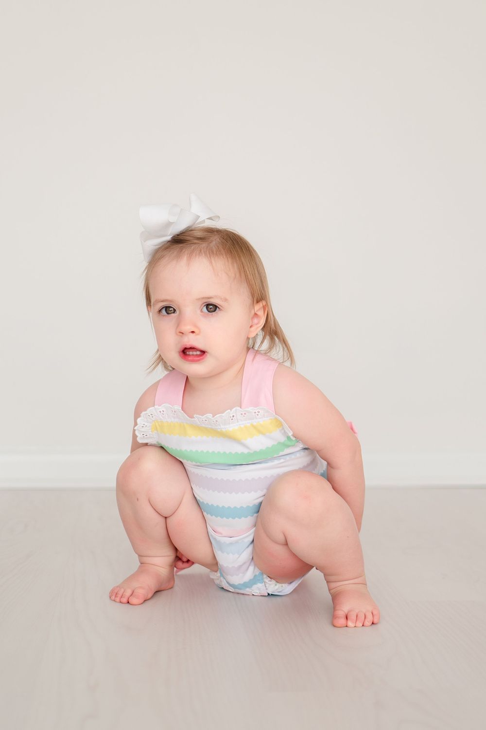 A baby in a pastel striped romper and white bow crouches on a light gray floor against a neutral background.