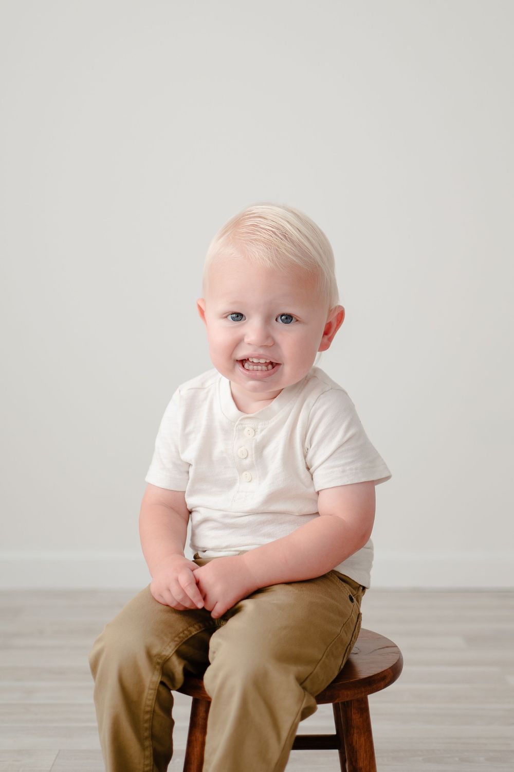 A young child wearing a white t-shirt and khaki pants sits smiling on a wooden stool against a light gray backdrop.