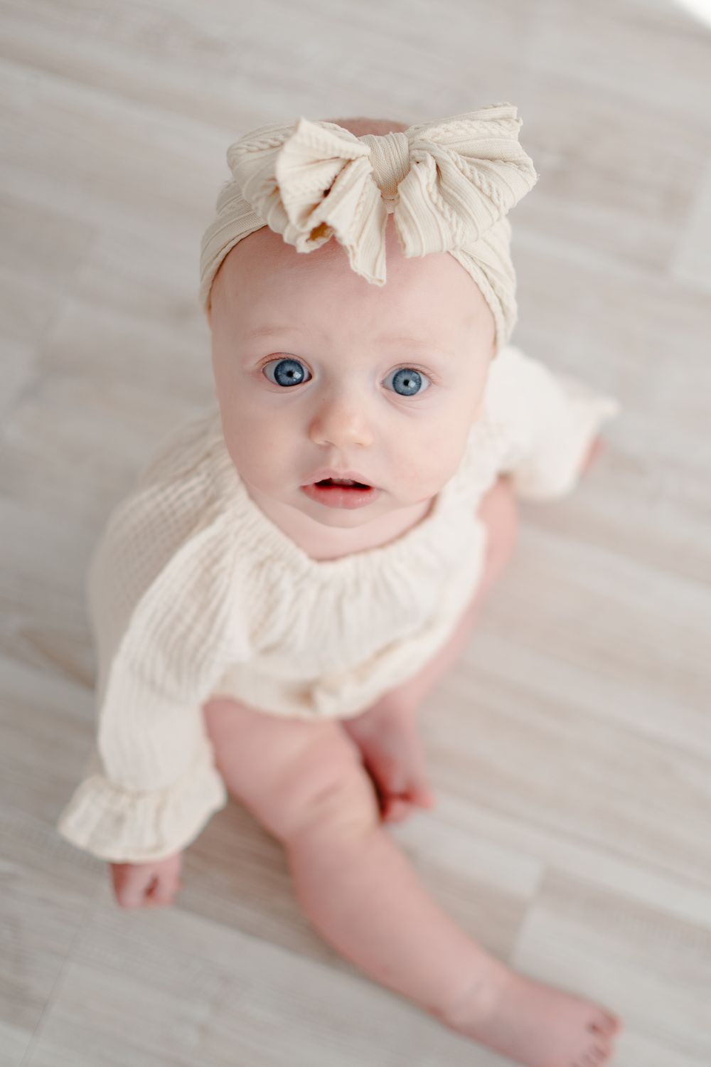 A small child in a cream-colored outfit with ruffles and a matching bow headband sits on a light wooden floor.