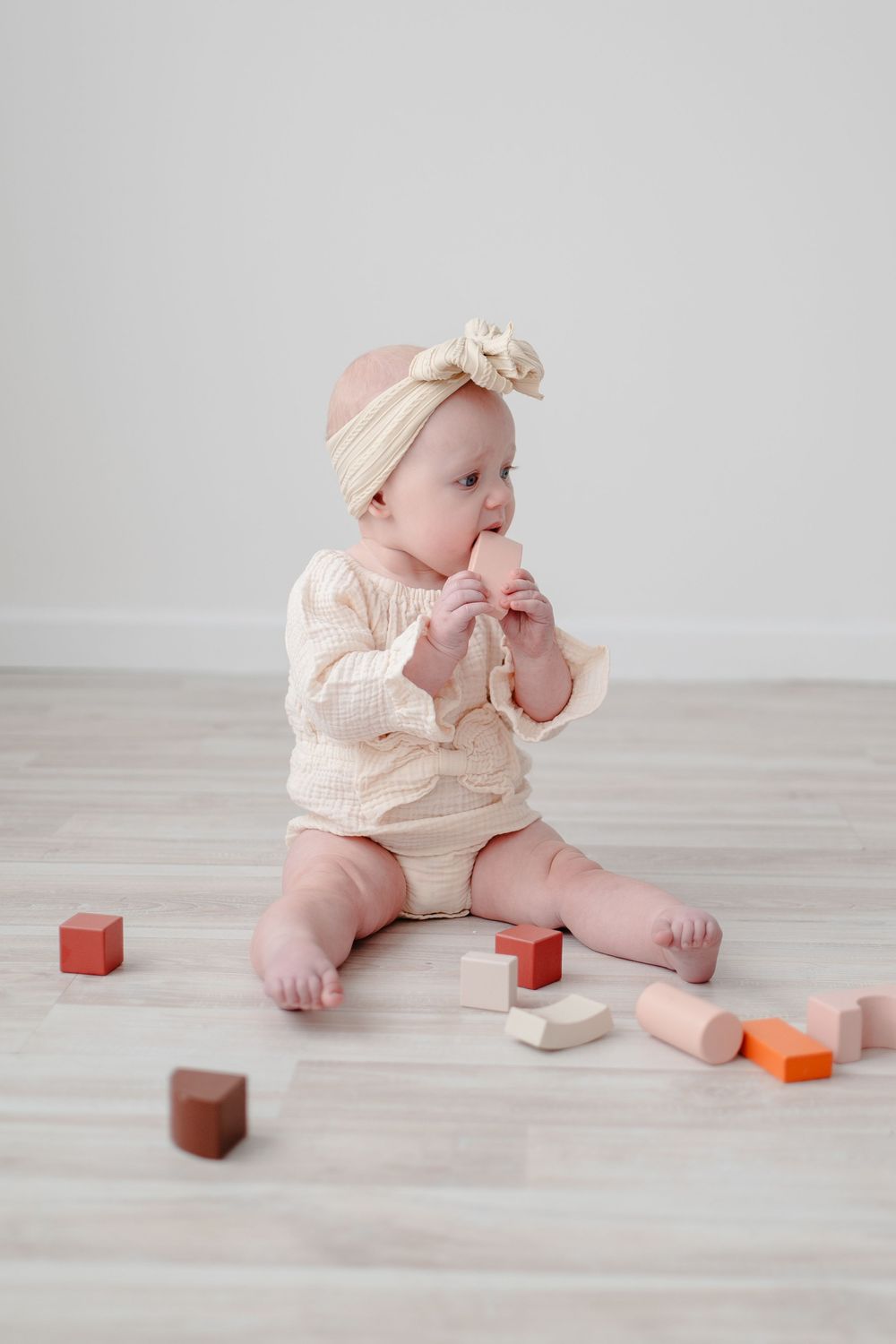 Baby in cream-colored outfit and bow headband sits on floor playing with wooden toy blocks on light wood flooring.