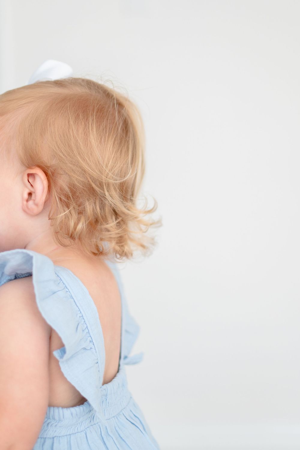 Young child with blonde curls wearing a light blue dress stands sideways against a white background.
