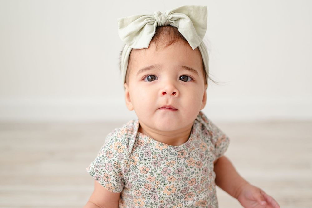 Baby wearing a floral print dress and cream bow headband sits on light wooden floor.