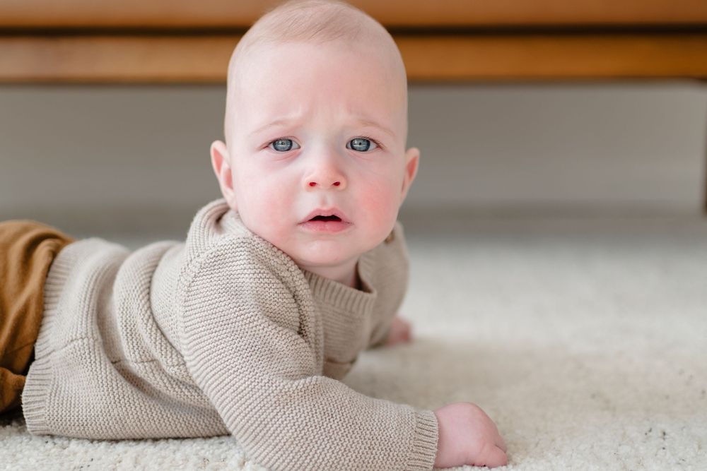 Baby wearing beige sweater lies on cream colored carpet while looking at camera.