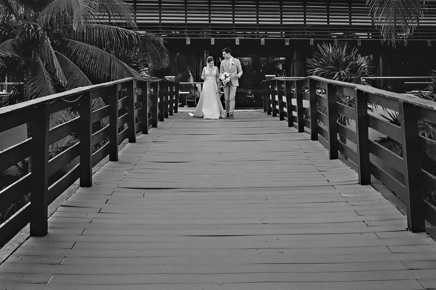 Black and white photo of a bride and groom walking away on a wooden boardwalk lined with railings and palm trees.