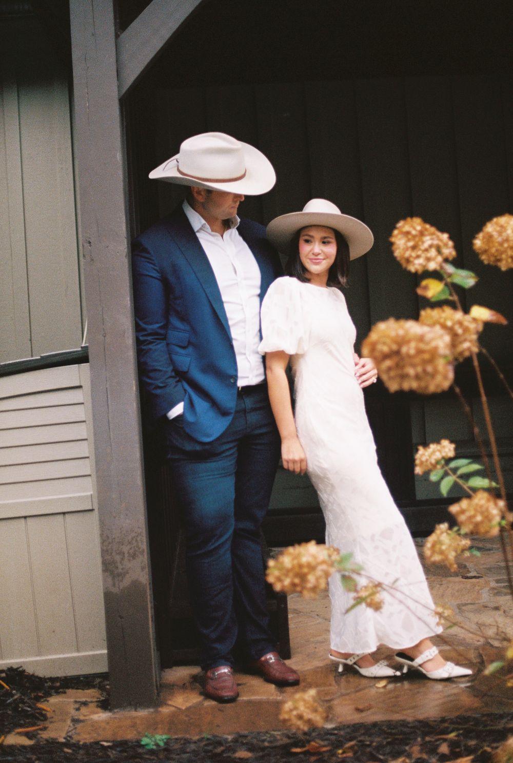 A couple in western attire poses together near dried hydrangeas and a rustic barn wall on a fall day.