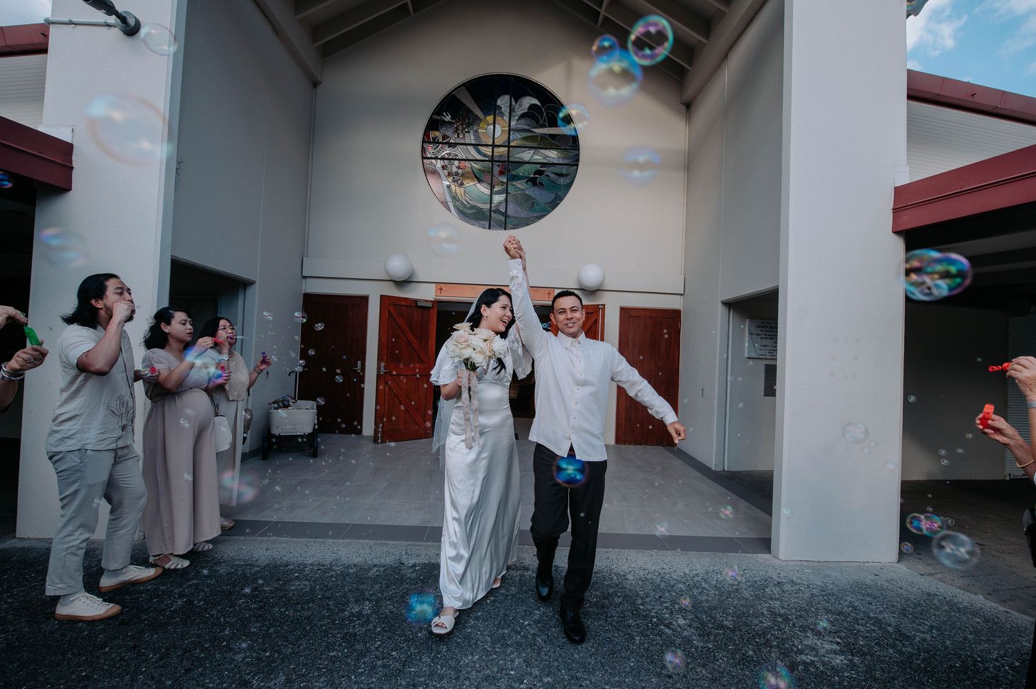 A couple exits their wedding venue with celebratory confetti while guests look on from behind in matching outfits.