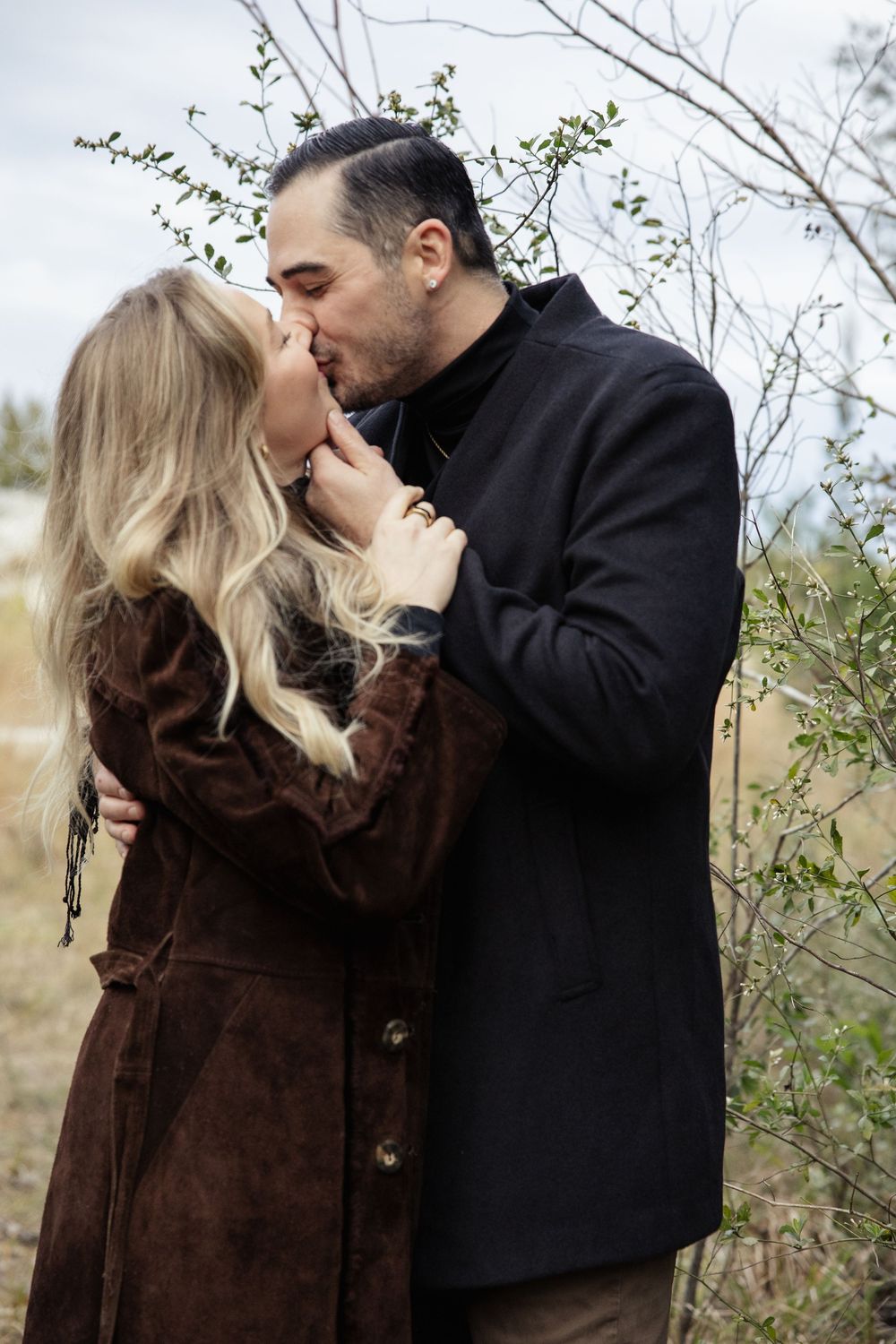 A romantic winter scene shows a couple sharing an intimate moment while wearing dark coats among bare tree branches.