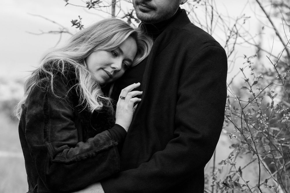 A couple embraces in a tender moment outdoors, with the woman resting her head against her partner's chest in black and white.