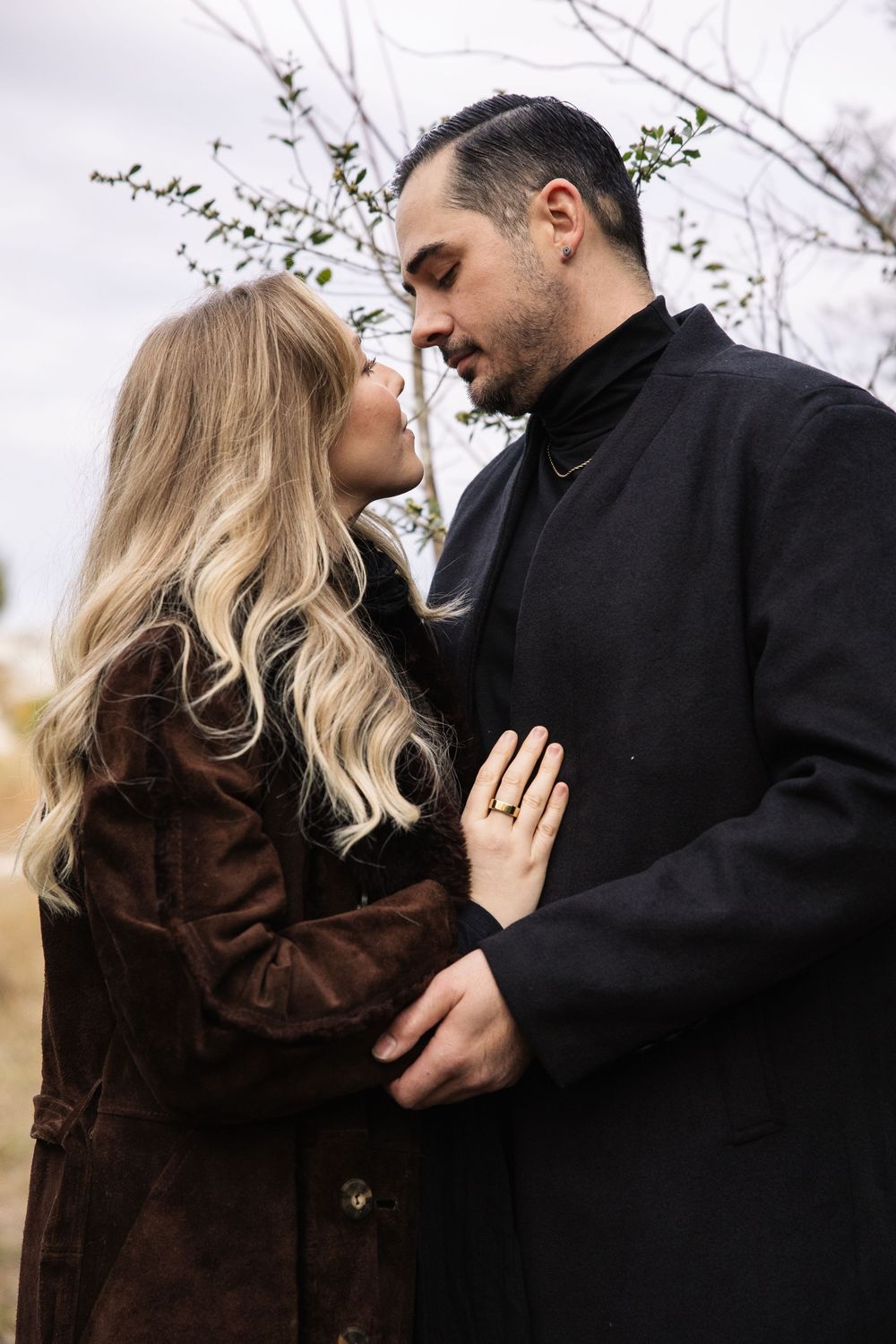 A romantic couple in winter coats sharing an intimate moment outdoors with bare trees in the background.