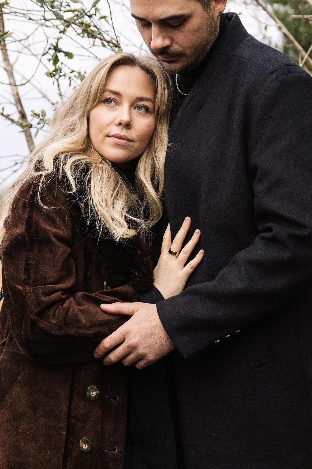 Romantic winter portrait of couple embracing outdoors, wearing dark coats against bare branches in moody natural lighting.