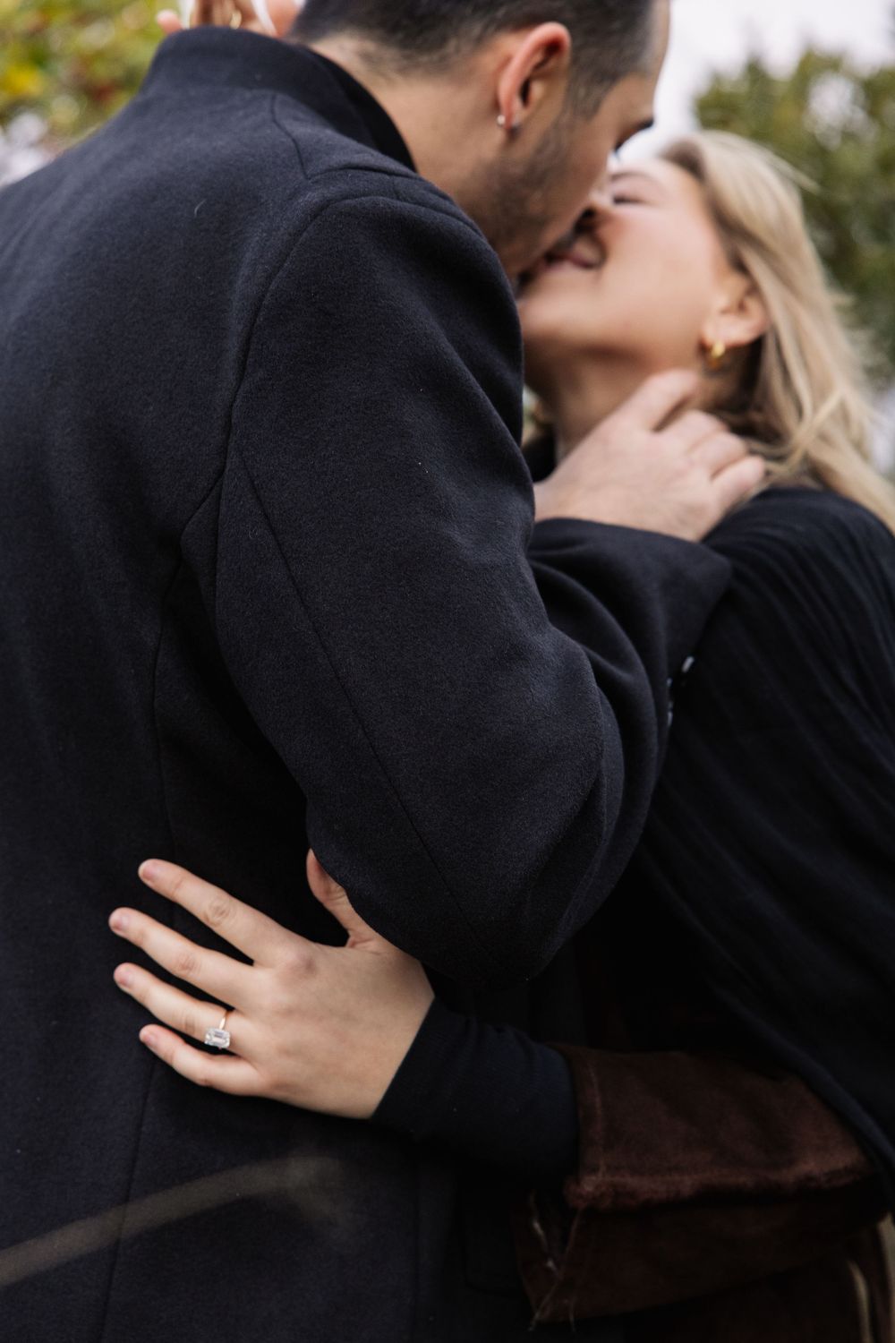A romantic couple in black coats share an intimate moment outdoors while an engagement ring sparkles on her hand.