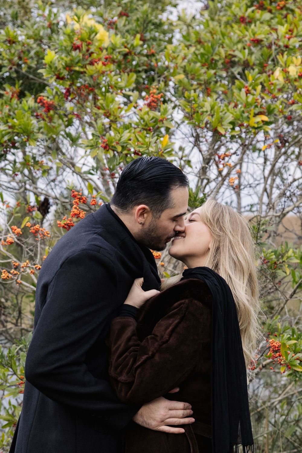 A couple in dark coats share a romantic kiss in front of autumn foliage with red berries in a garden setting.