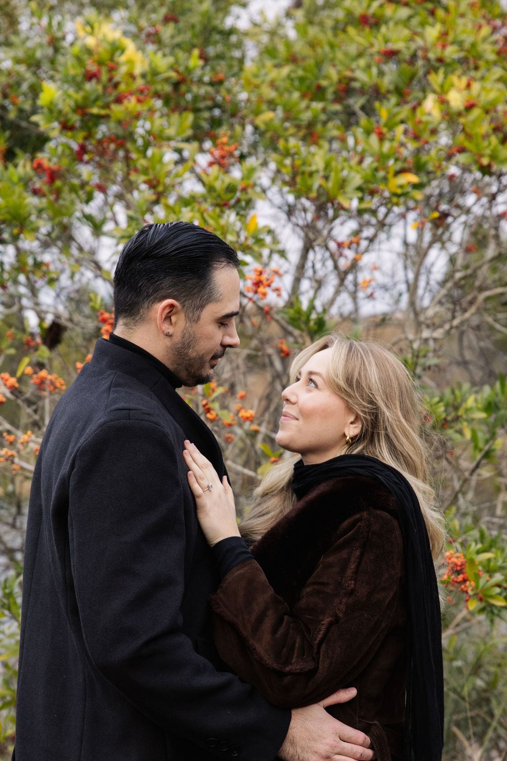 A romantic couple in winter coats embrace outdoors against autumn foliage and trees.
