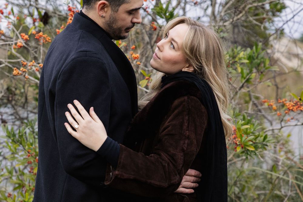 Couple in warm winter coats embrace outdoors near orange berries and foliage.