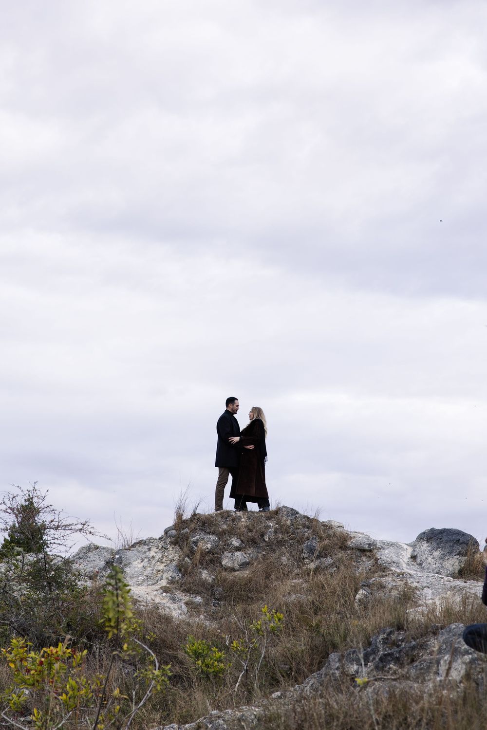 Two silhouetted figures stand close together on a rocky hilltop against an overcast sky with yellow wildflowers below.