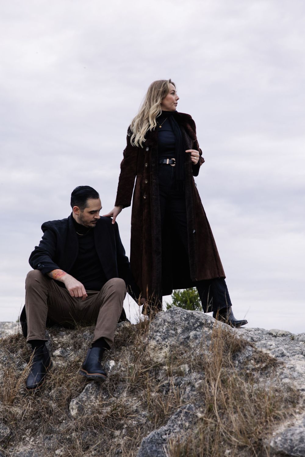 Two people in dark winter coats pose dramatically on rocky terrain against an overcast sky.