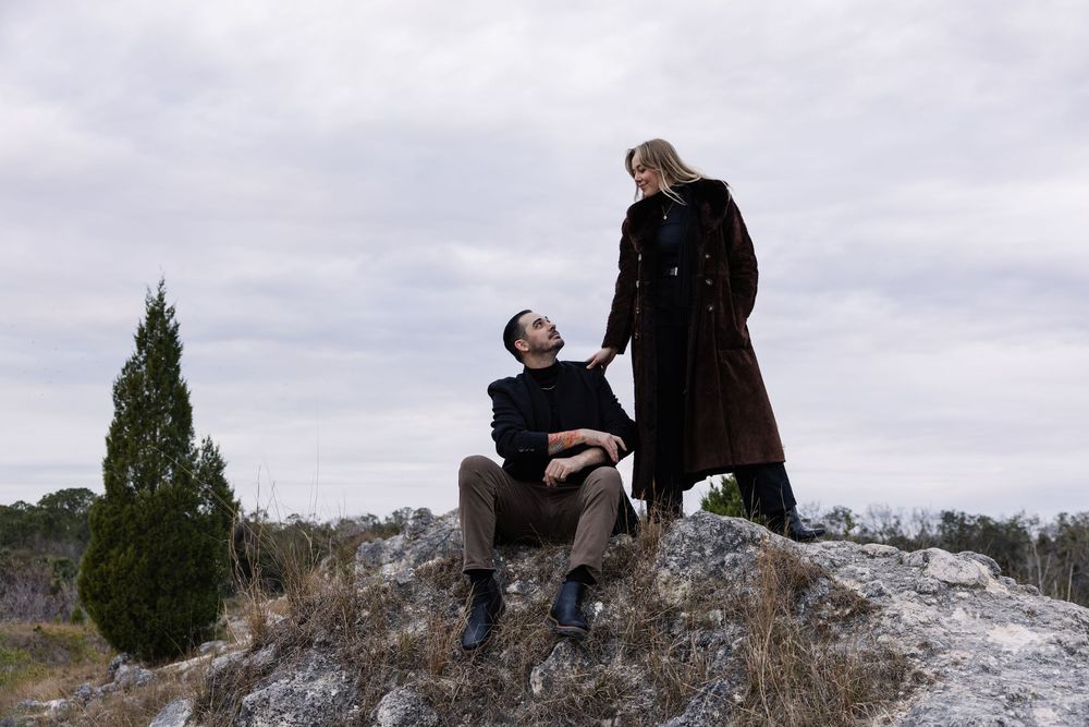 Two people stand together on a rocky outcrop under cloudy skies during what appears to be a proposal moment.