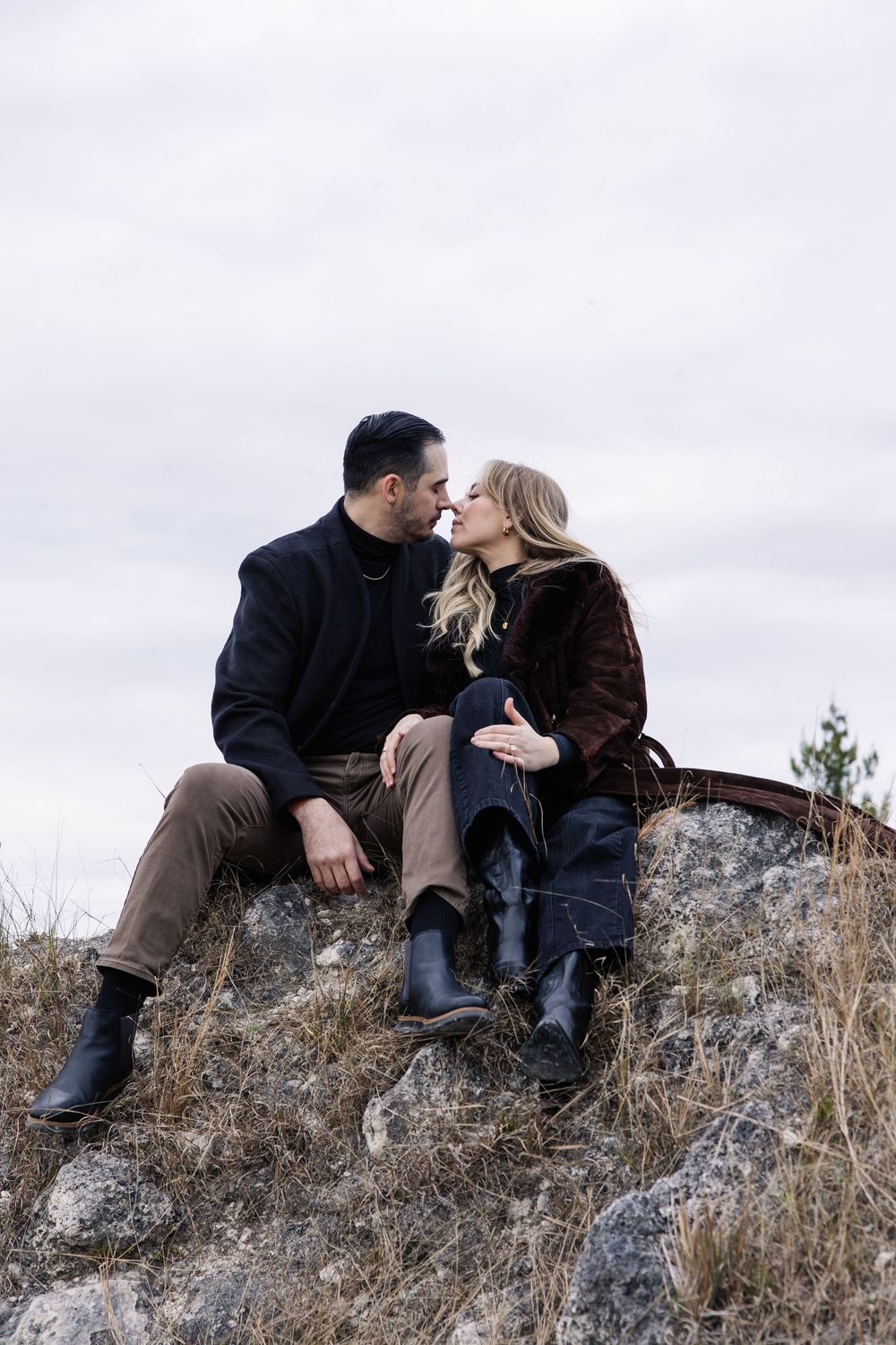 A romantic couple sits on a rocky hillside sharing an intimate moment while wearing winter coats against a cloudy sky.