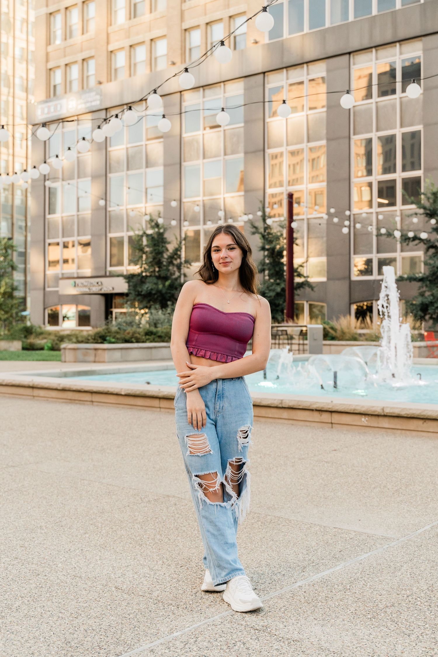 Perrysburg High School senior in burgundy top and distressed jeans standing near building with string lights and fountain at golden hour.