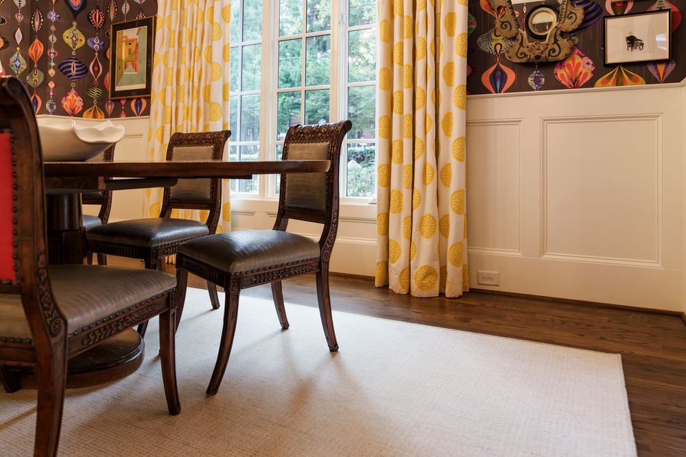 Elegant dining room with dark wood chairs, yellow curtains, and patterned wallpaper features hardwood floors and wainscoting.
