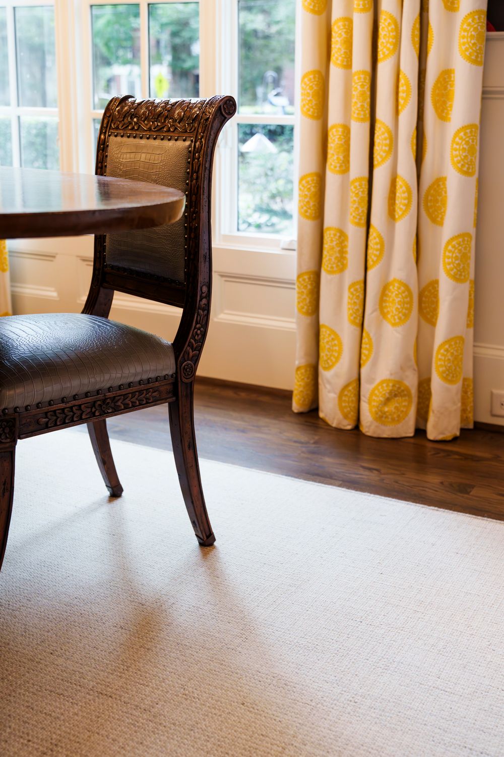 A dark wooden accent chair sits against yellow patterned curtains on beige carpeting in a bright room.