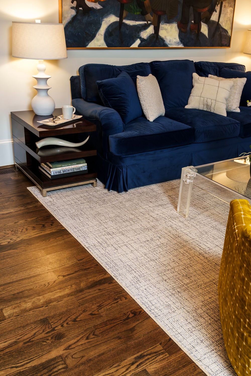 Modern living room features a navy blue sofa, white area rug, hardwood floors and yellow glass accent table.