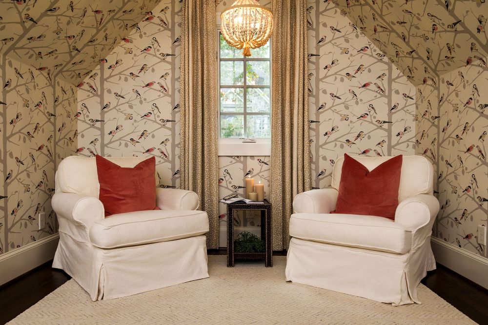 Cozy reading nook with two white slipcovered chairs, red accent pillows, and bird-patterned wallpaper beside a tall window.