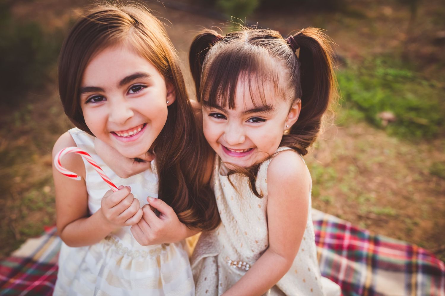 Two young girls in white dresses laughing and embracing while sitting outdoors in a natural setting.